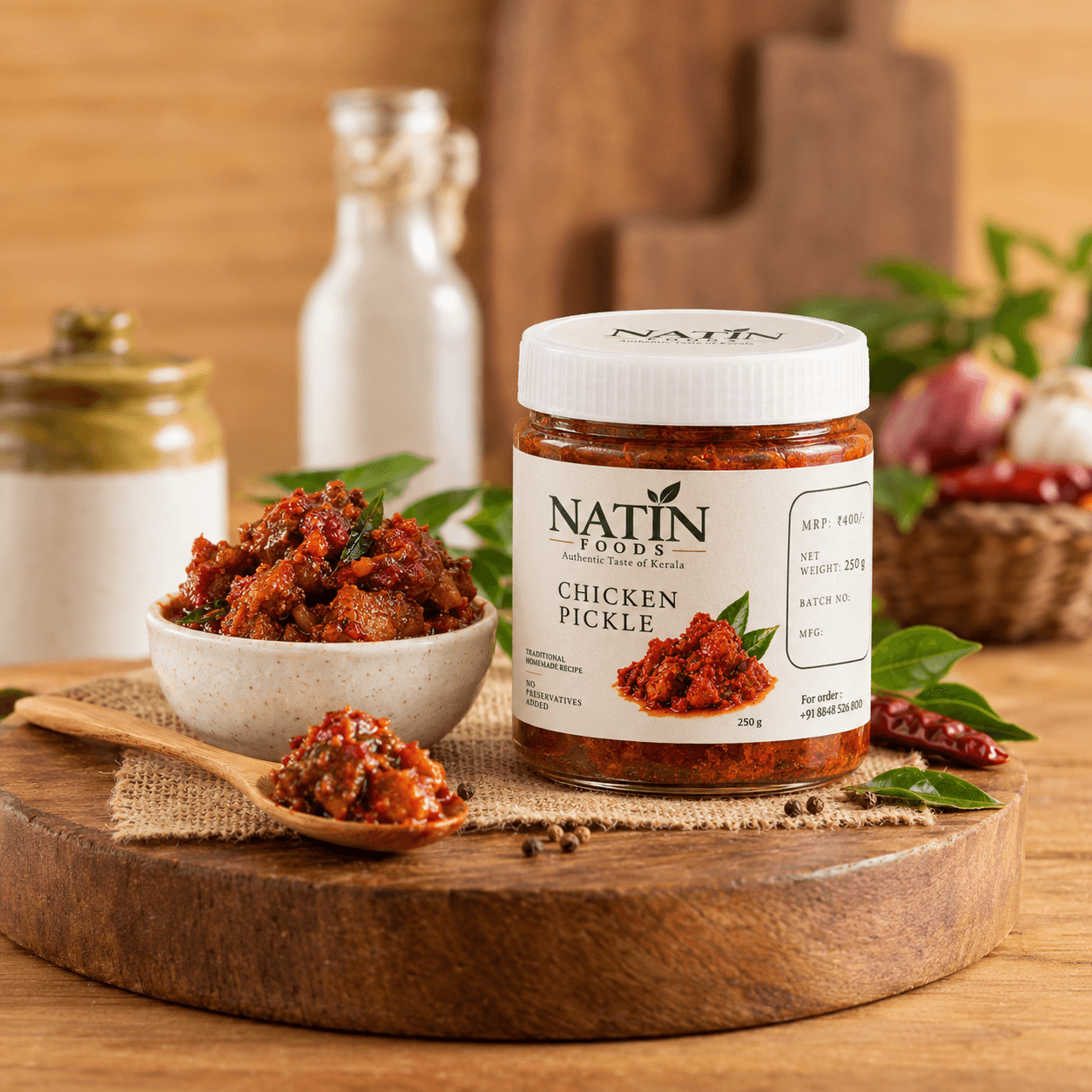 Glass jar of Natin Foods chicken pickle with a white cap and printed 250 g label, beside a stone bowl of pickle, a wooden spoon and curry leaves on a board